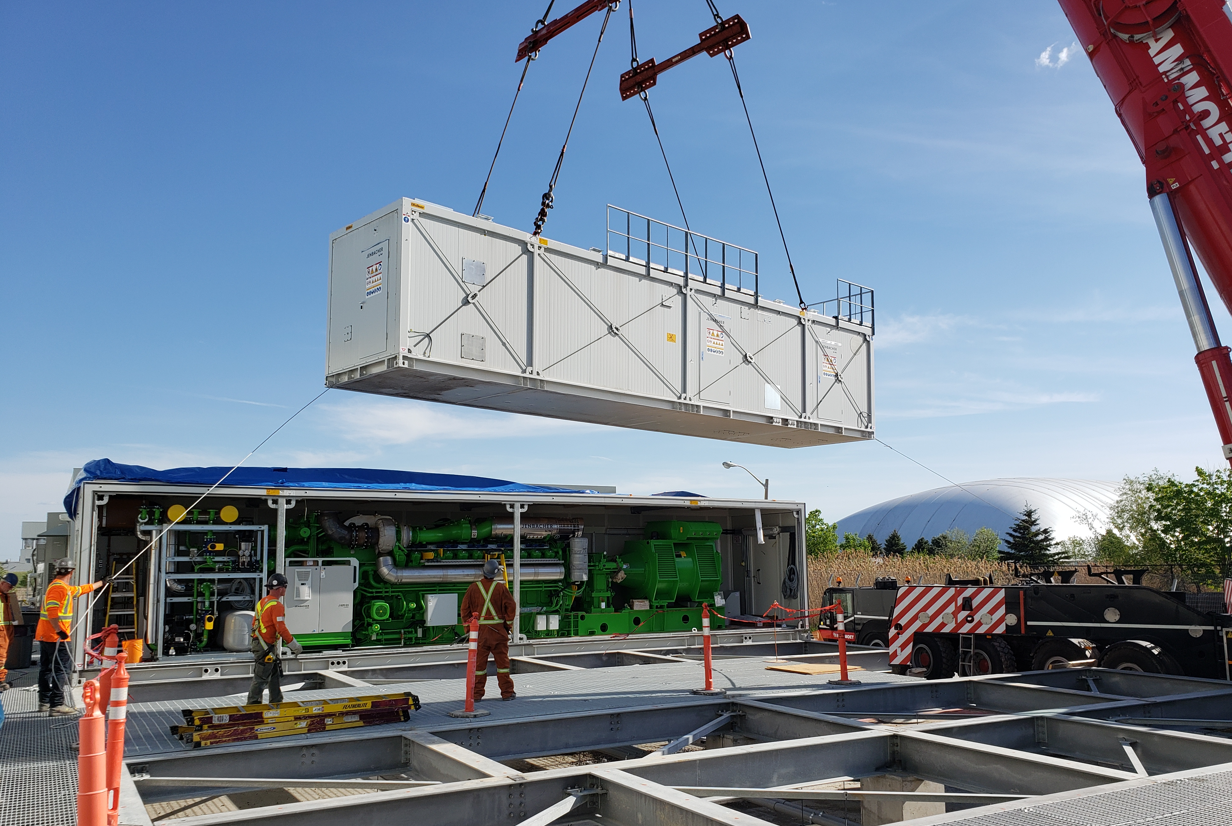 Workers operate crane to lift large industrial container at construction site, with equipment and structure visible below.