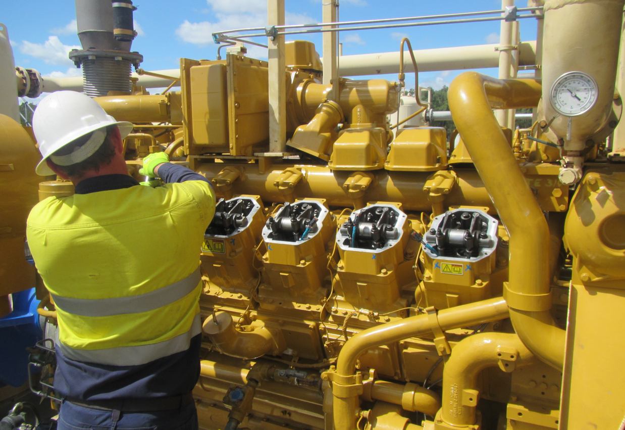 Technician in a high-visibility jacket working on an industrial machine engine outdoors under a blue sky.