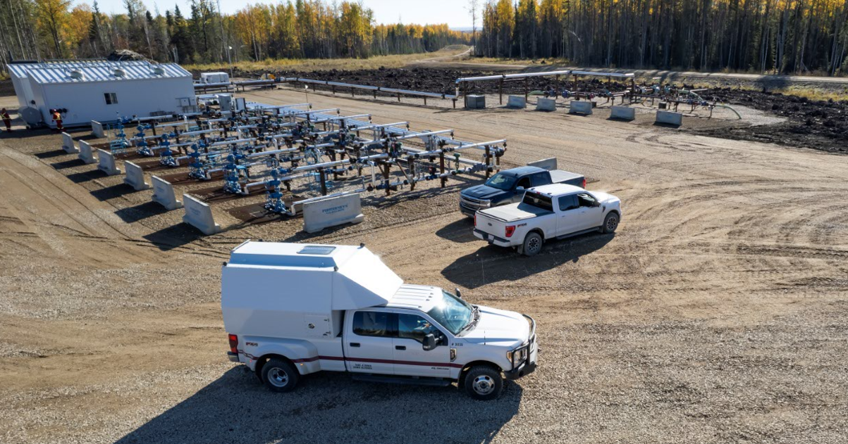 Oil and gas field facility with white service trucks, pipelines, and forest background in a clear day.