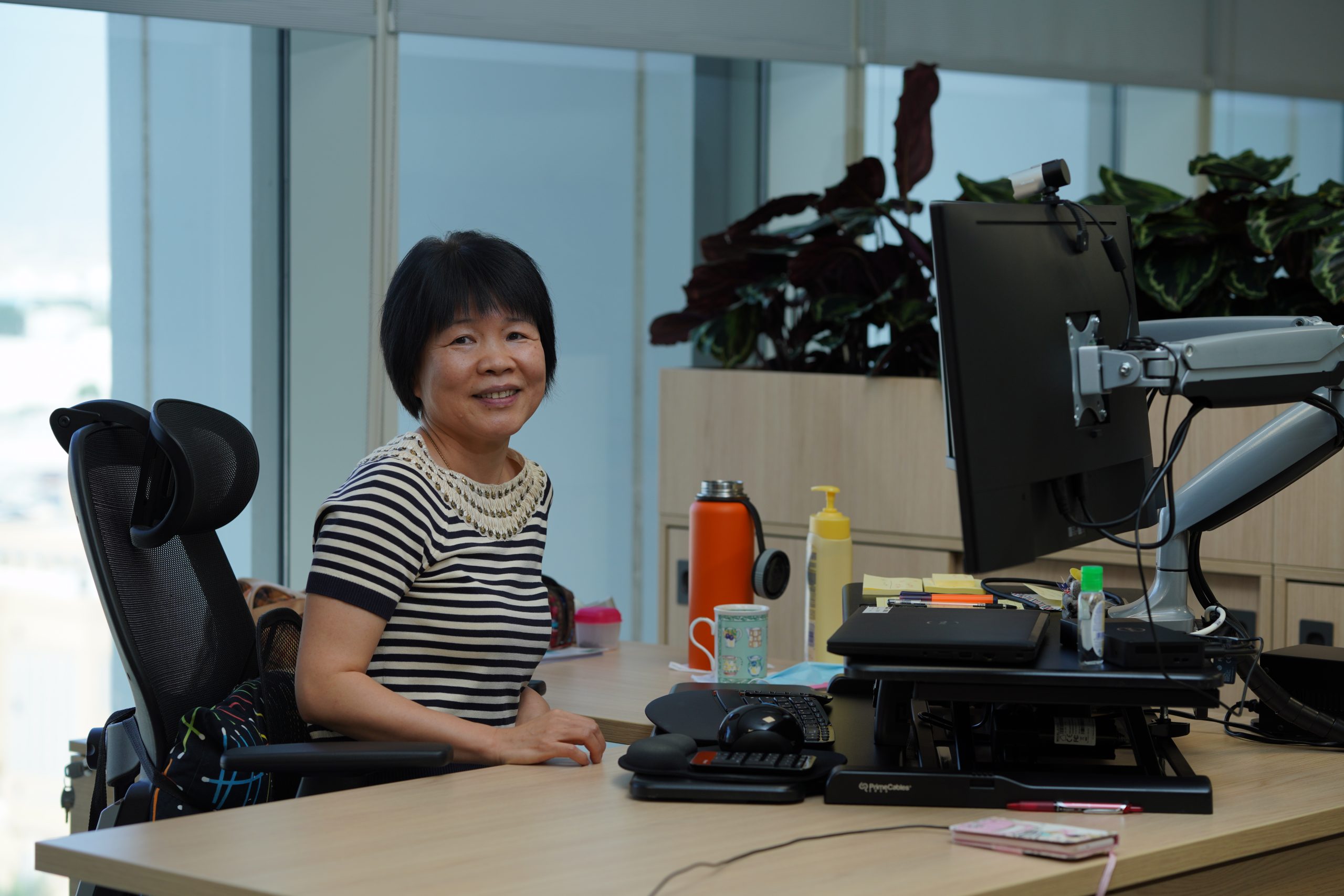 Smiling woman sits at an office desk with a computer monitor, plants in background, near large windows, and office supplies on desk.