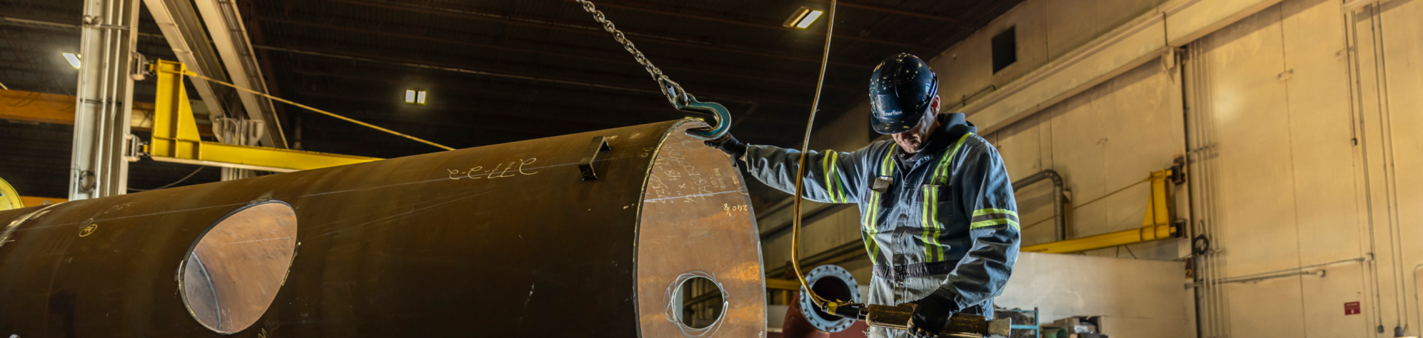 Worker in safety gear inspecting large industrial metal cylinder inside a factory setting with overhead crane.