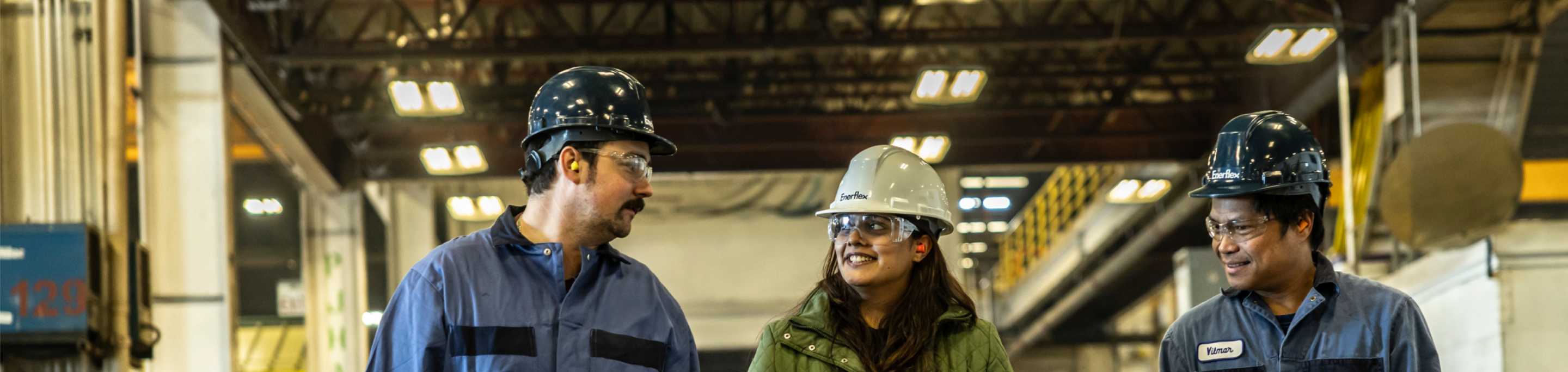 Industrial workers in hard hats and safety glasses having a conversation in a factory, showcasing teamwork and safety.