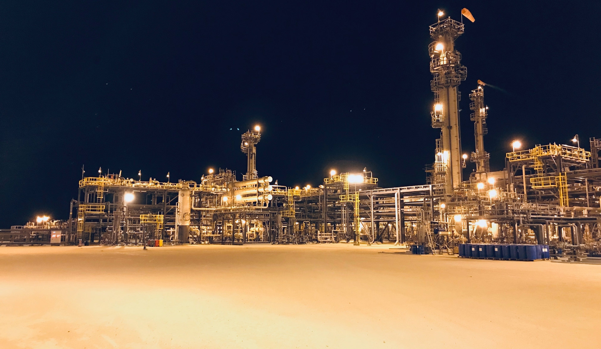 Night view of an illuminated industrial oil refinery with machinery and pipelines against a dark sky backdrop.