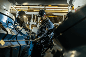 Two industrial workers in hard hats and safety gear inspecting machinery in a factory, ensuring proper operation.