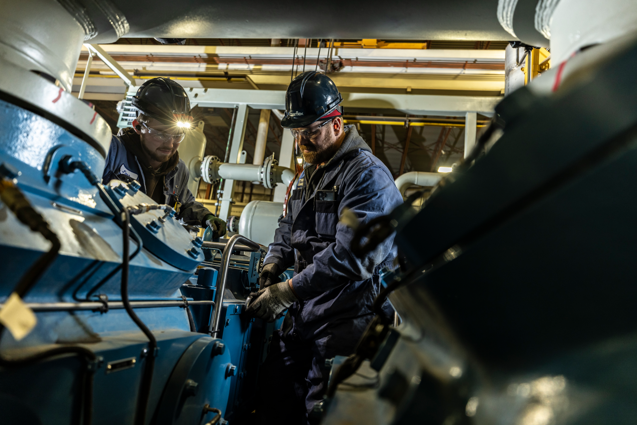 Two industrial workers in hard hats and safety gear inspecting machinery in a factory, ensuring proper operation.