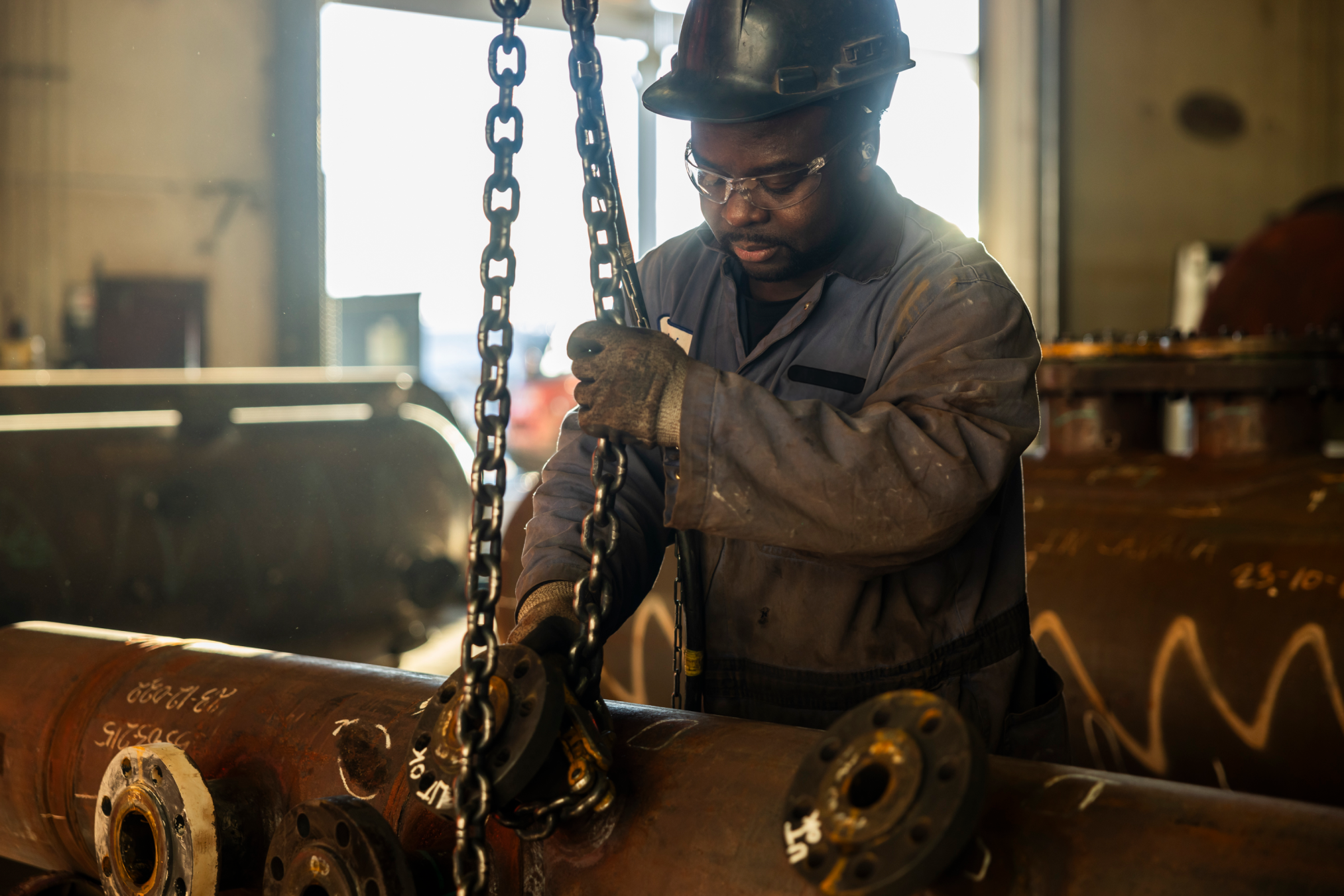 Industrial worker in a hard hat operates heavy machinery with chains in a factory, focusing on large metal pipes.