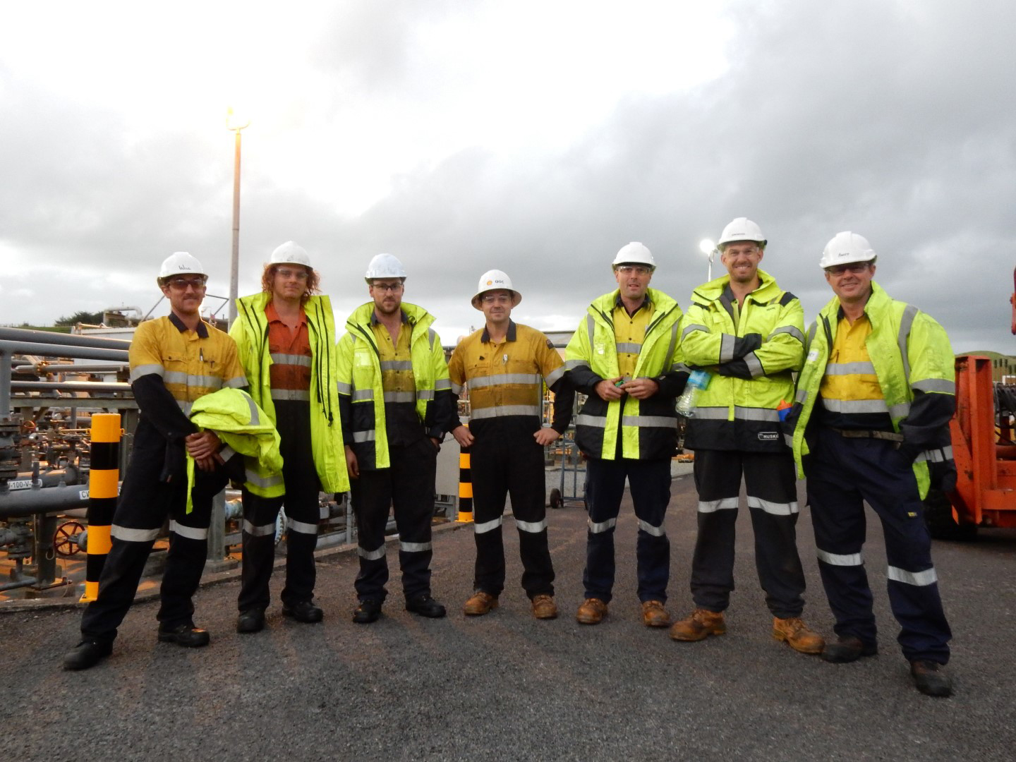 Construction workers in safety gear standing together at an industrial site under cloudy sky.