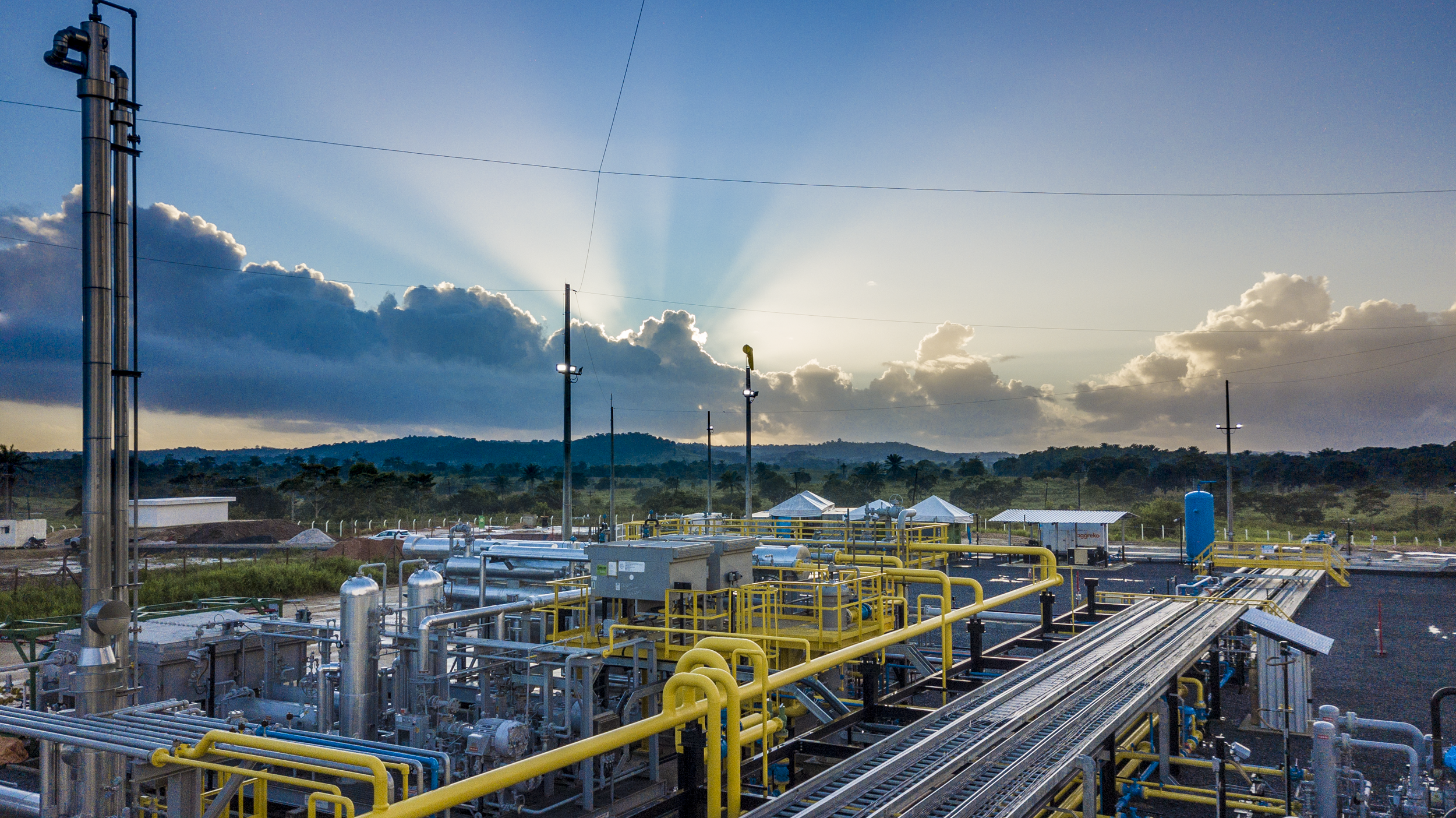 Industrial complex at sunset, equipped with pipelines and machinery, against a backdrop of hills and dramatic clouds.