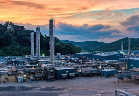 Industrial facility with tall structures and pipelines set against a colorful sunset sky, nestled in a green hilly area.